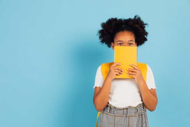 Colegiala afroamericana divertida que cubre su rostro con un libro amarillo sobre fondo azul concepto de regreso a la escuela