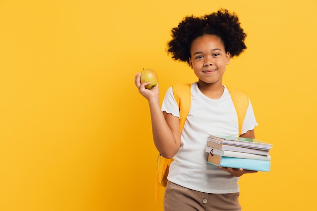 Colegiala afroamericana feliz sosteniendo libros de cuadernos y manzana de almuerzo en el espacio de copia de fondo amarillo