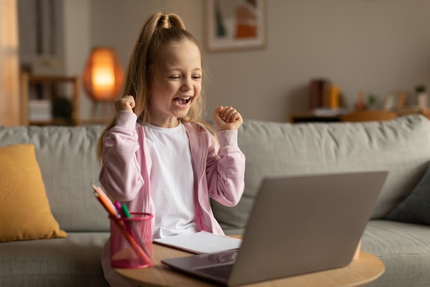 Colegiala alegre en la computadora portátil agitando los puños en casa