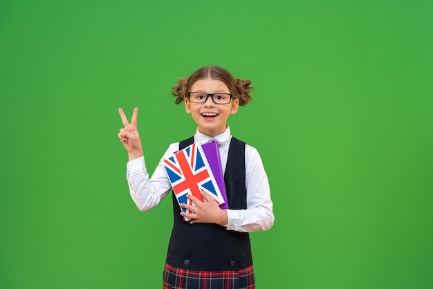 una colegiala alegre con un libro de texto de inglés en uniforme escolar con corbata. El alumno muestra dos dedos sobre un fondo verde aislado y copia el espacio. aprendiendo idiomas extranjeros,