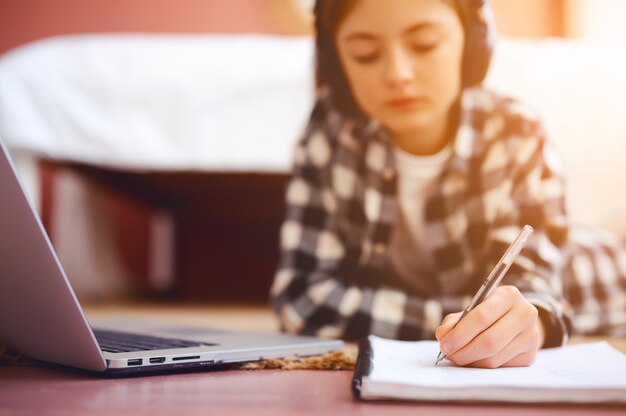 Colegiala con auriculares escribiendo en el cuaderno cerca de la computadora portátil