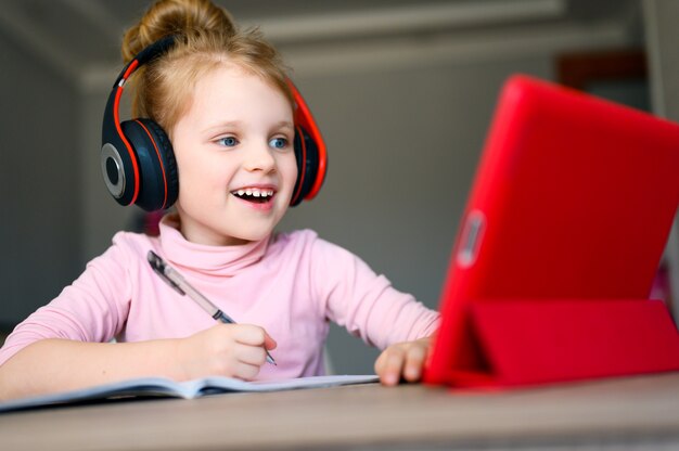 Colegiala en auriculares estudiando en casa con tableta digital, sonriendo y haciendo la tarea escolar.