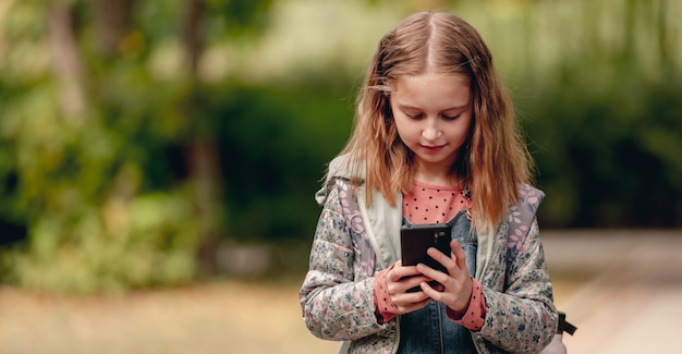 Colegiala bonita con smartphone al aire libre. Linda niña de pie con teléfono celular en el parque otoño