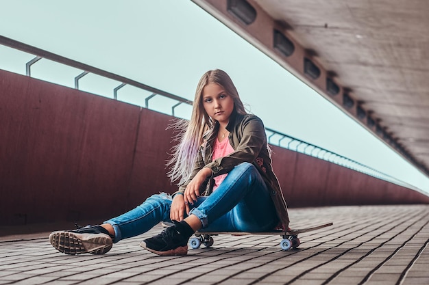 Colegiala con cabello rubio vestida con ropa de moda sentada en una patineta en un puente peatonal.