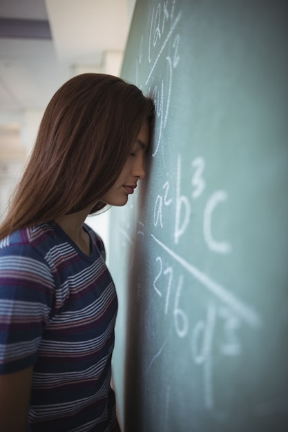 Colegiala cansada de pie con los ojos cerrados cerca de pizarra en el aula