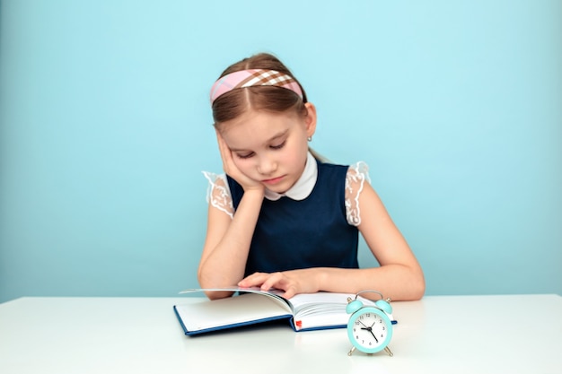 Colegiala cansada que estudia con un libro y un reloj sobre fondo azul claro.