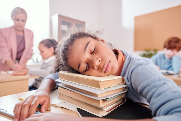 Foto colegiala cansada en ropa casual manteniendo la cabeza en la pila de libros mientras toma una siesta junto al escritorio en la lección