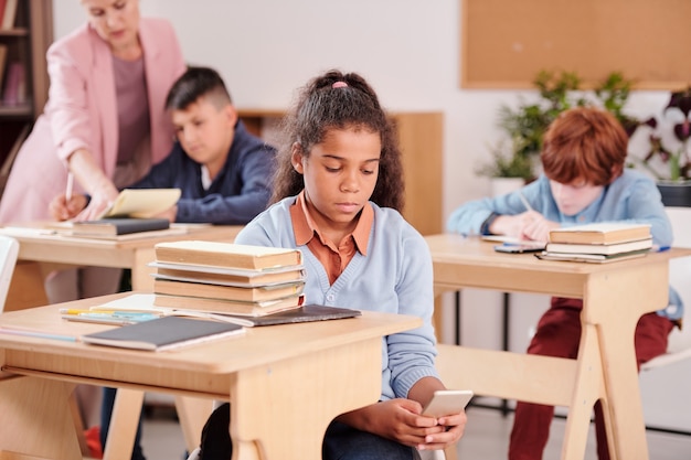 Colegiala contemporánea de raza mixta que usa un teléfono inteligente y lo oculta mientras busca la respuesta correcta en la red durante el trabajo individual en la lección