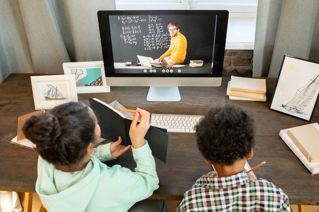 Colegiala diligente mostrando notas en copybok al maestro en el monitor de la computadora mientras está sentado en la mesa junto a su hermano durante la lección en línea