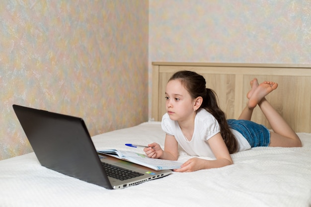 Colegiala estudiando en casa con una tableta portátil portátil y haciendo la tarea escolar en la cama con libros de capacitación