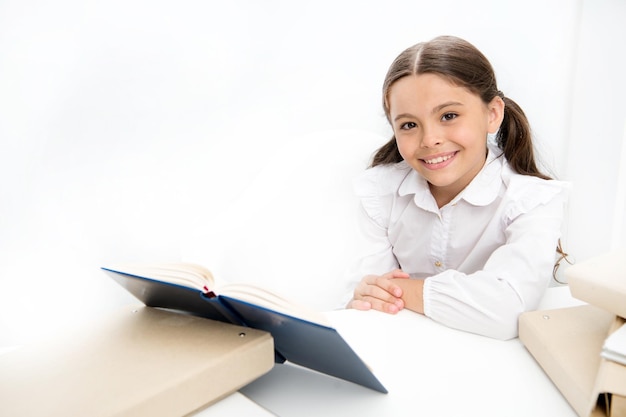 Colegiala estudiando libro de texto Niño uniforme escolar cara feliz leer libro Emocionado por el conocimiento Concepto de educación en el hogar Libro interesante para niños Niña leer libro sentarse mesa interior blanco