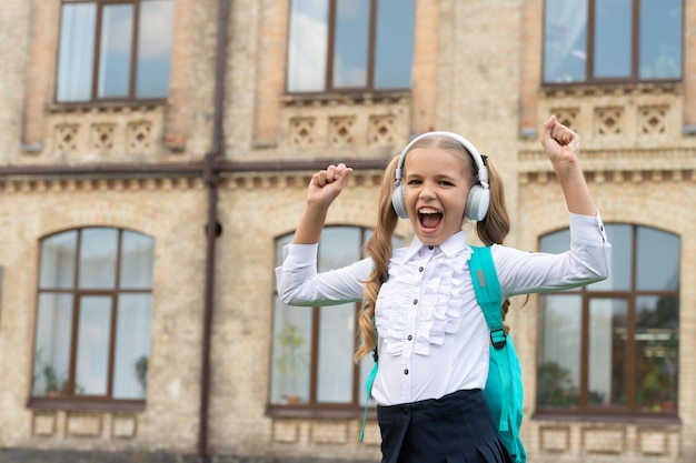 Colegiala feliz en uniforme haciendo gesto ganador escuchando música en auriculares