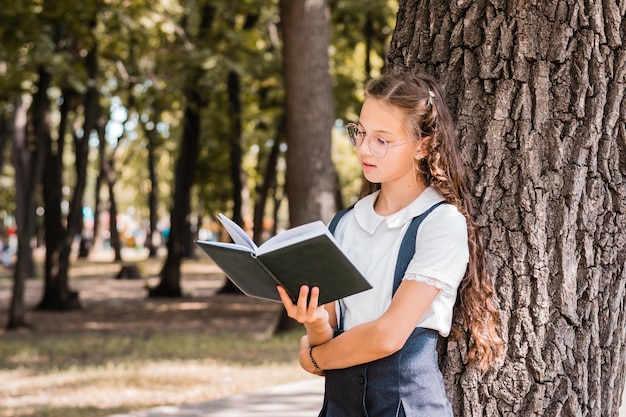 Colegiala con gafas lee un libro cerca de un árbol en el parque