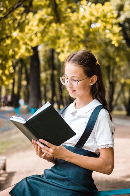 Colegiala con gafas leyendo un libro en un día cálido en el parque Vista vertical