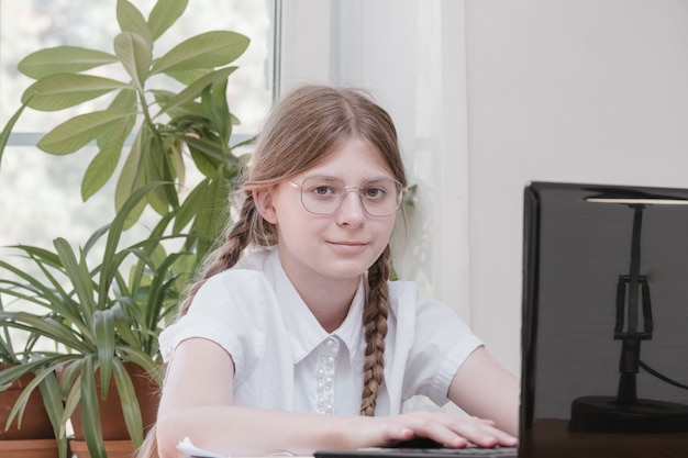 Una colegiala inteligente hace la tarea sentada en su escritorio, escribe con un bolígrafo en sus notas de escritura y usa una computadora portátil. Ella está aprendiendo el idioma en línea, haciendo tareas escolares en casa, escuchando conferencias