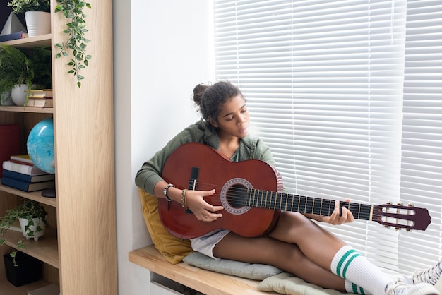 Colegiala intercultural relajada tocando la guitarra mientras está sentado en el alféizar de la ventana en el entorno hogareño