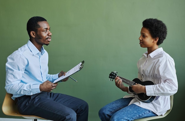 Colegiala joven feliz con ukelele consultando con su profesor en la lección de música mientras ambos se sientan