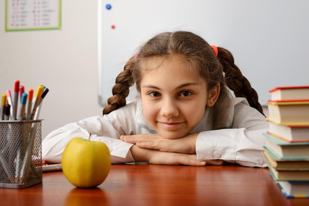 Foto colegiala juguetona y alegre sentada en el escritorio con libros, lápices y una manzana durante la lección