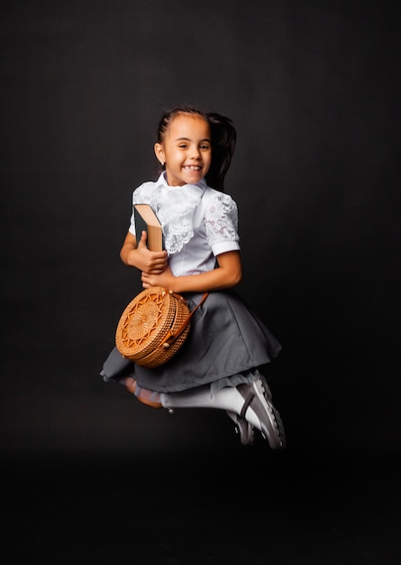 Foto colegiala linda alegre sostiene un libro y una bolsa redonda y salta sobre un fondo oscuro.