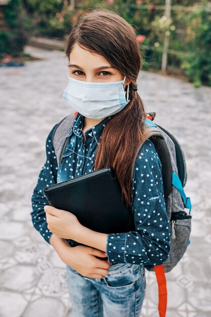 Foto colegiala con una máscara médica en el rostro y una computadora portátil en la mano
