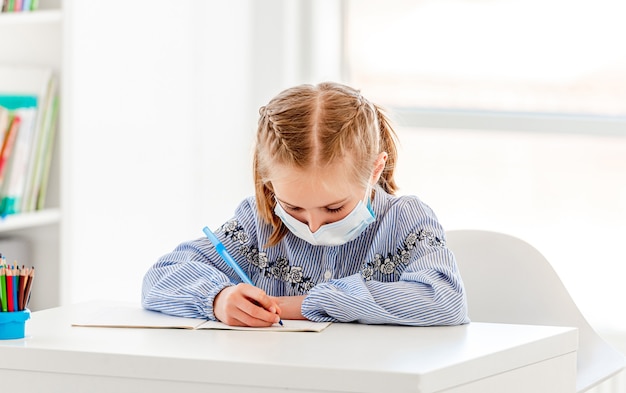 Colegiala en máscara protectora escribiendo con lápiz en el cuaderno sentado en el pupitre de la escuela