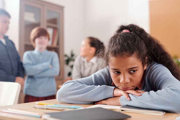 Colegiala molesta u ofendida sentada junto al escritorio y manteniendo la barbilla y las manos en el cuaderno