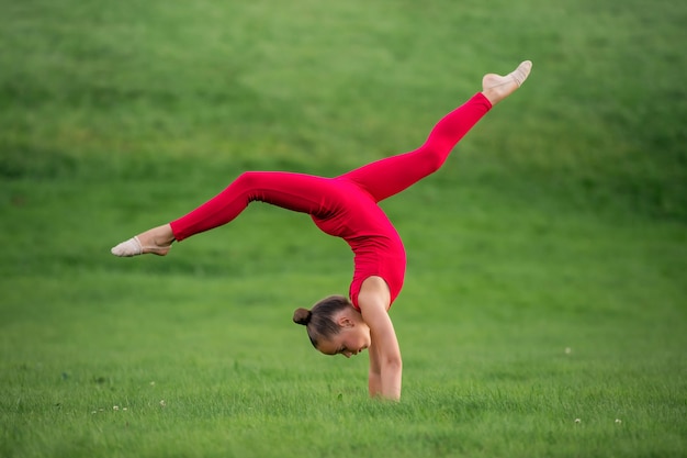 Colegiala con un mono rojo brillante se dedica a la gimnasia sobre el césped, haciendo ejercicios de estiramiento