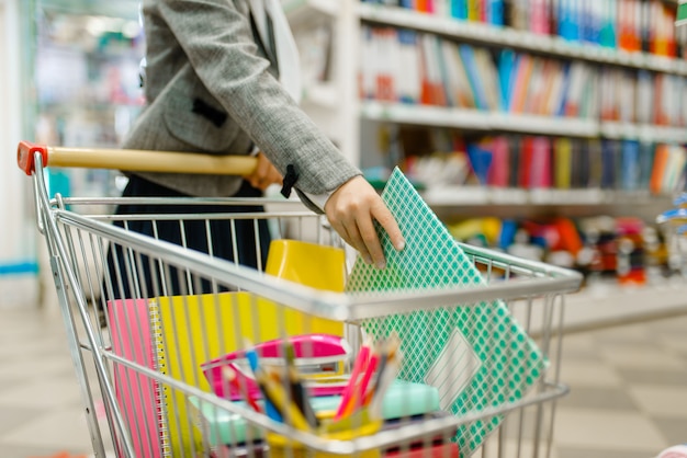 La colegiala pone un cuaderno en el carro en el estante de la papelería. Niña comprando suministros de oficina en la tienda, escolares en el supermercado