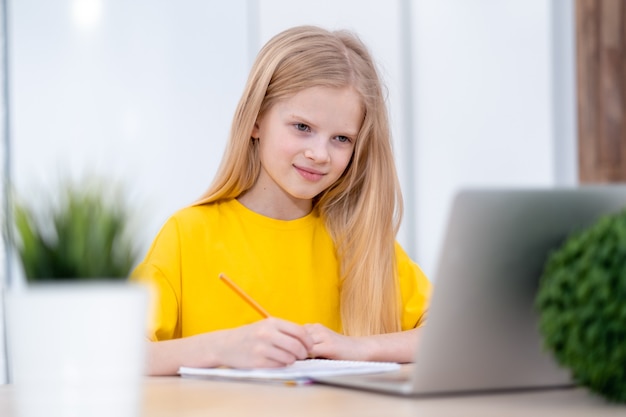 Colegiala rubia estudiando en casa haciendo los deberes escolares con el aprendizaje a distancia en línea portátil