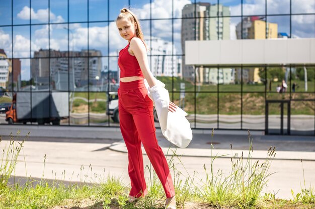 Colegiala rubia joven en un traje rojo y una chaqueta blanca posando en el estacionamiento