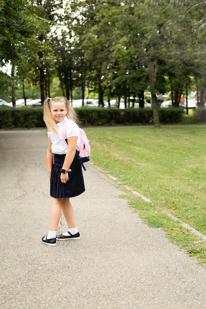 Colegiala rubia sonriente en uniforme escolar con mochila rosa yendo a la escuela al aire libre