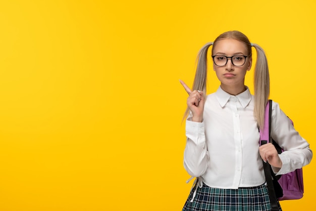 Colegiala seria del día mundial del libro con cola de caballo señalando con el dedo con mochila