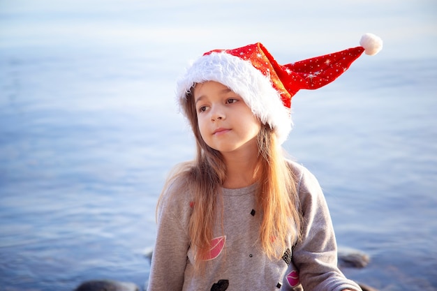 Una colegiala con un sombrero de Año Nuevo se sienta en la orilla del mar. Navidad en países cálidos. Vacaciones y vacaciones de año nuevo en el mar.