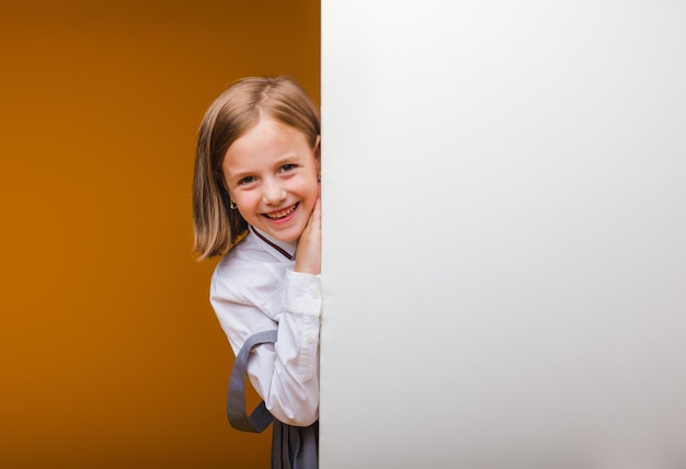 Una colegiala sonriente se para detrás de un panel blanco en blanco resaltado en un fondo amarillo Un espacio vacío para el texto se asoma desde detrás de la pancarta