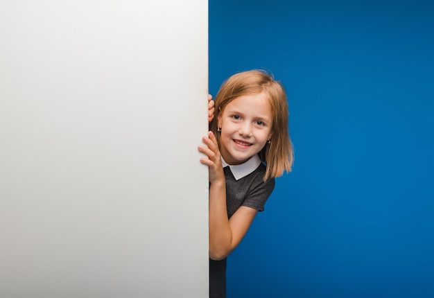 Una colegiala sonriente se encuentra detrás de un panel blanco en blanco resaltado en un fondo azul. Un espacio vacío para el texto se asoma desde detrás de la pancarta.