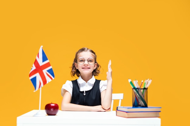 Colegiala sonriente levantando la mano sentada en el escritorio durante la lección de inglés lección bandera de gran bretaña