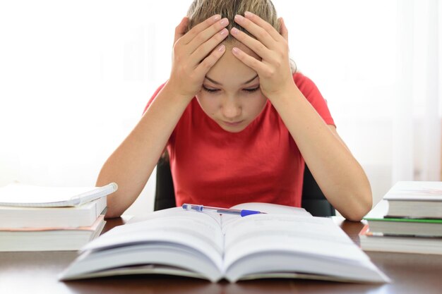 La colegiala sostiene su cabeza en sus manos sobre una pila de libros en el escritorio y está cansada y frustrada con el problema de la tarea en la escuela