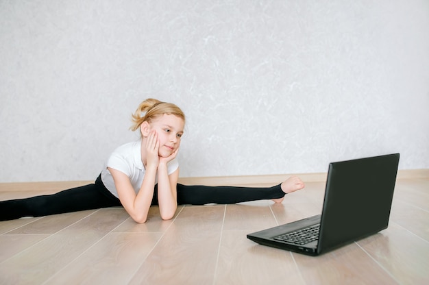 Colegiala viendo videos en línea en la computadora portátil y haciendo ejercicios deportivos: yoga, gimnasia, coreografía. Quédate en casa. Concepto de autoaislamiento, cuarentena, educación en línea.