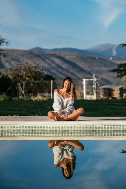 Foto colha uma mulher descalça sentada de pernas cruzadas perto da piscina com água reflexiva calma e sorrindo no quintal