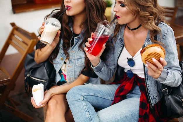 Colheita de garotas deslumbrantes em roupas casuais, bebendo café e limonada e comendo hambúrguer com batatas fritas no café.