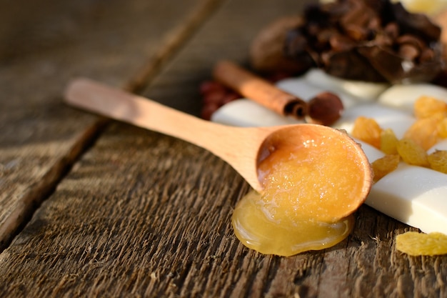 Foto colher de pau com mel em uma mesa de madeira ao lado de telhas de chocolate branco, passas, paus de canela