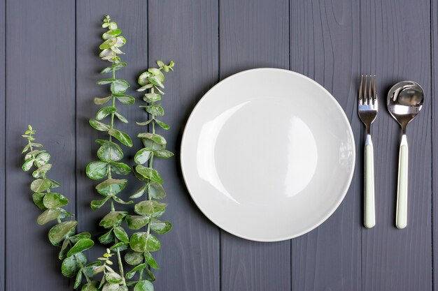 Colher, garfo, placa cinza e raminho de eucalipto verde na mesa de madeira cinza