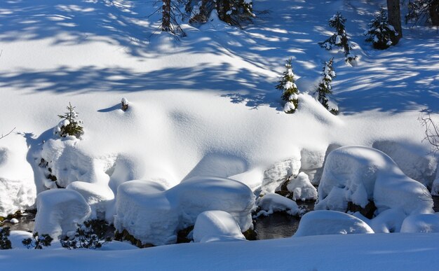 Colina de montanha de neve de inverno com pequenos pinheiros e riacho sob nevascas.