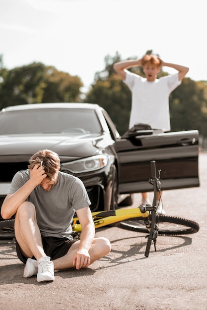 Colisão na estrada. Cara em roupa esportiva caindo da bicicleta e tocando a cabeça e um motorista ruivo empolgado de camiseta branca perto de um carro aberto