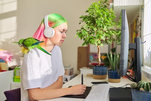 Foto college-student, attraktives teenager-mädchen mit kopfhörern zu hause, am computer mit grafiktablett studierend, mit grünem papagei auf der schulter sitzend