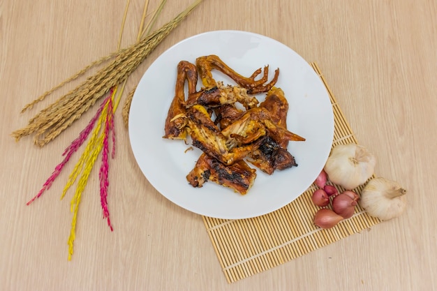 Se coloca pollo asado en un plato blanco sobre la mesa y hay ajos, cebollas y espigas de arroz, por ahí.