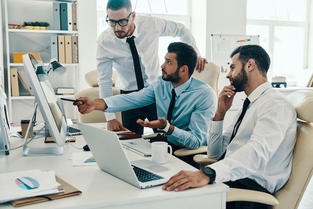 Colocando ideias em algo real. Grupo de jovens homens modernos em trajes formais, trabalhando usando computadores enquanto estão sentados no escritório