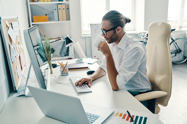 Colocando ideias em algo real. Jovem pensativo em uma camisa, trabalhando usando o computador e bebendo café enquanto está sentado no escritório