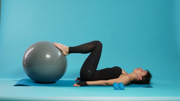 Colocar a un adulto usando una pelota tonificante de fitness para entrenar los músculos de las piernas frente a la cámara. Mujer haciendo ejercicio físico y entrenamiento corporal con equipo de gimnasia en colchoneta de yoga, entrenando en estudio.