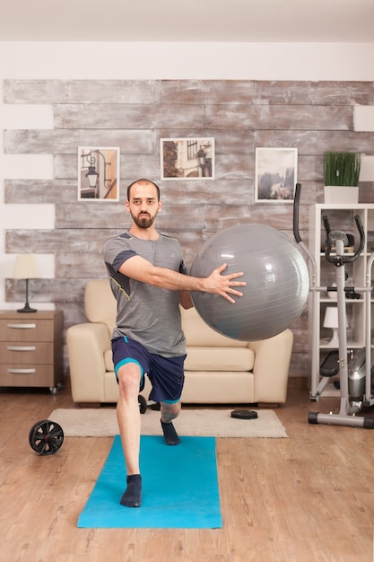 Colocar al hombre haciendo estocadas en la estera de yoga con pelota suiza durante la pandemia mundial.